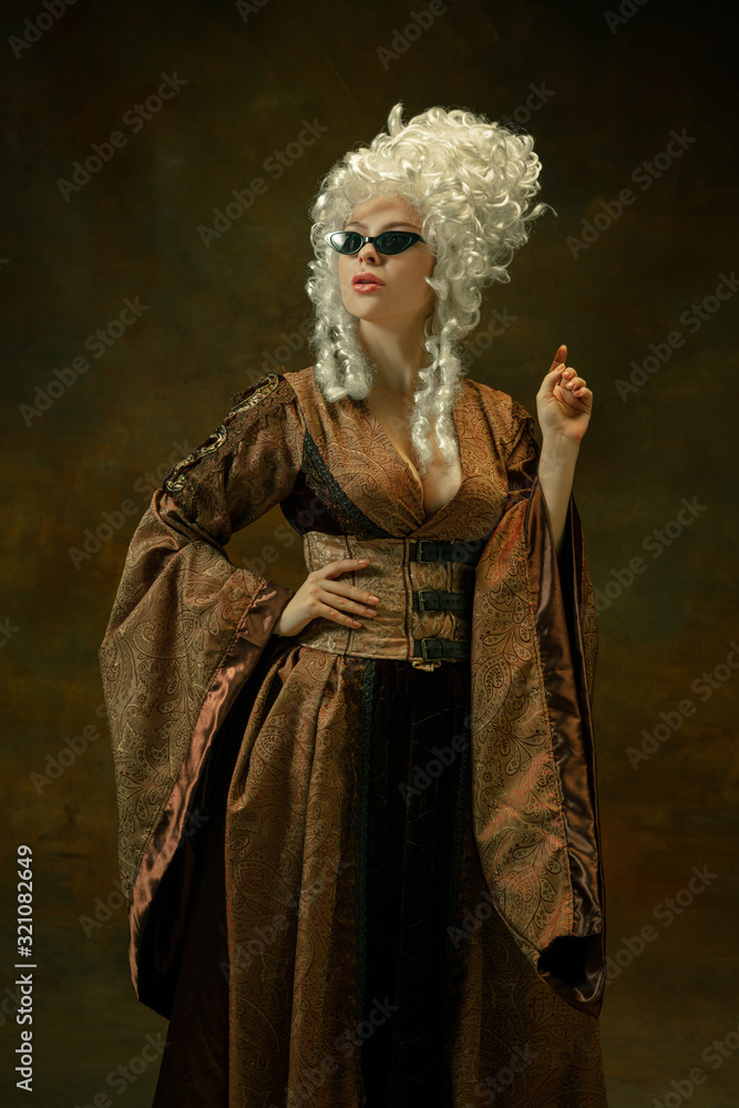 Trying on new eyewear. Portrait of medieval young woman in brown vintage clothing on dark background. Female model as a duchess, royal person. Concept of comparison of eras, modern, fashion, beauty.