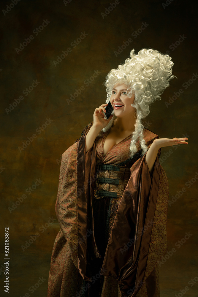 Talking on phone. Portrait of medieval young woman in brown vintage clothing on dark background. Female model as a duchess, royal person. Concept of comparison of eras, modern, fashion, beauty.