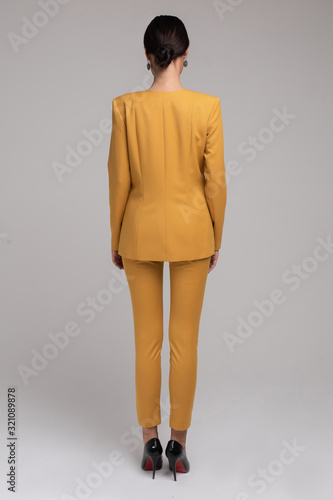 Back view of faceless blonde woman with hairstyle wearing skinny trousers and green jackets with heels over white background.