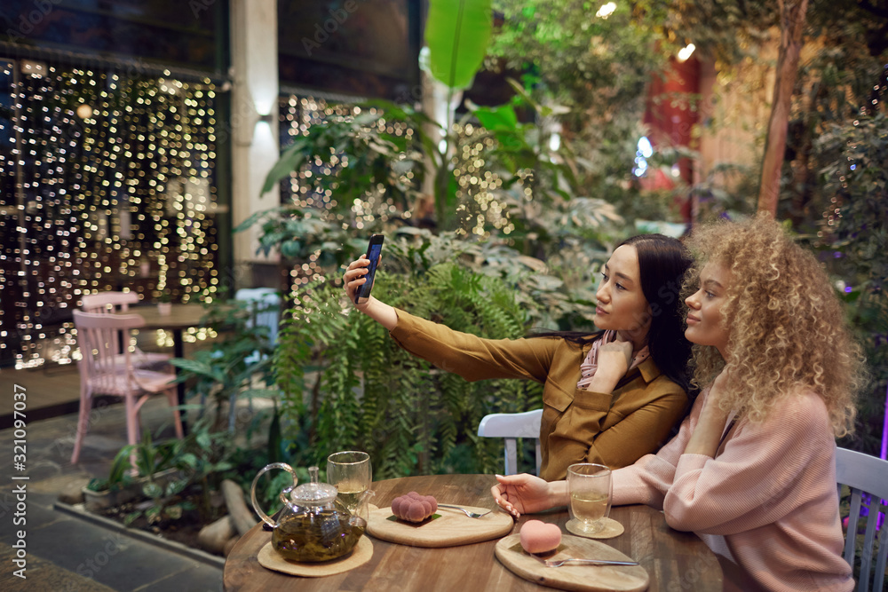 Asian young woman holding her mobile phone and making selfie portrait with her friend while they drinking tea in cafe