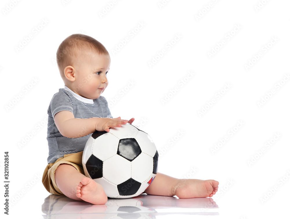 Infant baby boy toddler in striped t-shirt and pants is sitting holding football soccer ball playing on white Infant baby boy toddler in striped t-shirt and pants is sitting holding football soccer ball playing on white