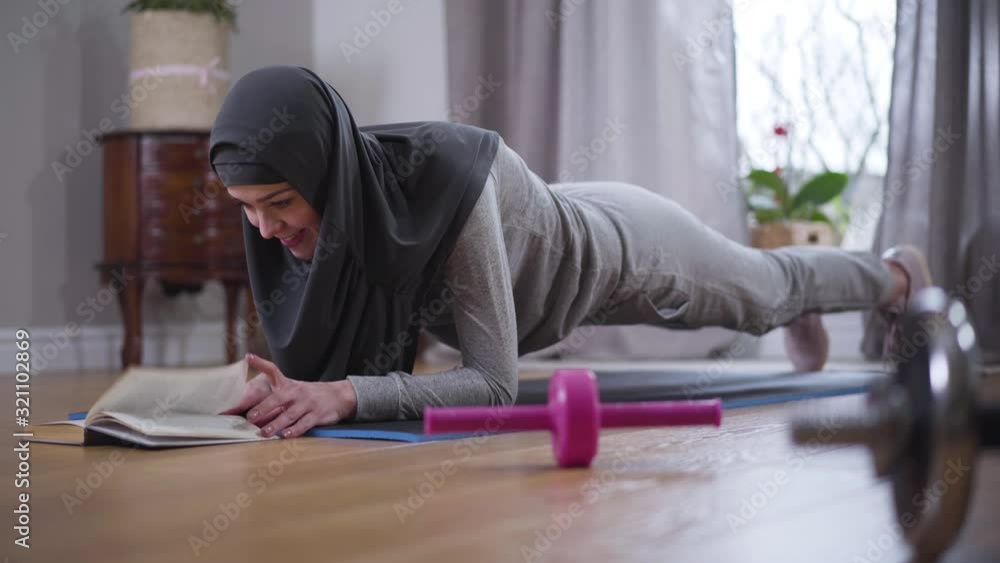 Strong Muslim woman in hijab and sportswear doing sport planc exercise and reading book. Confident young woman practicing sport and hobby at home. Leisure, lifestyle, enjoyment.