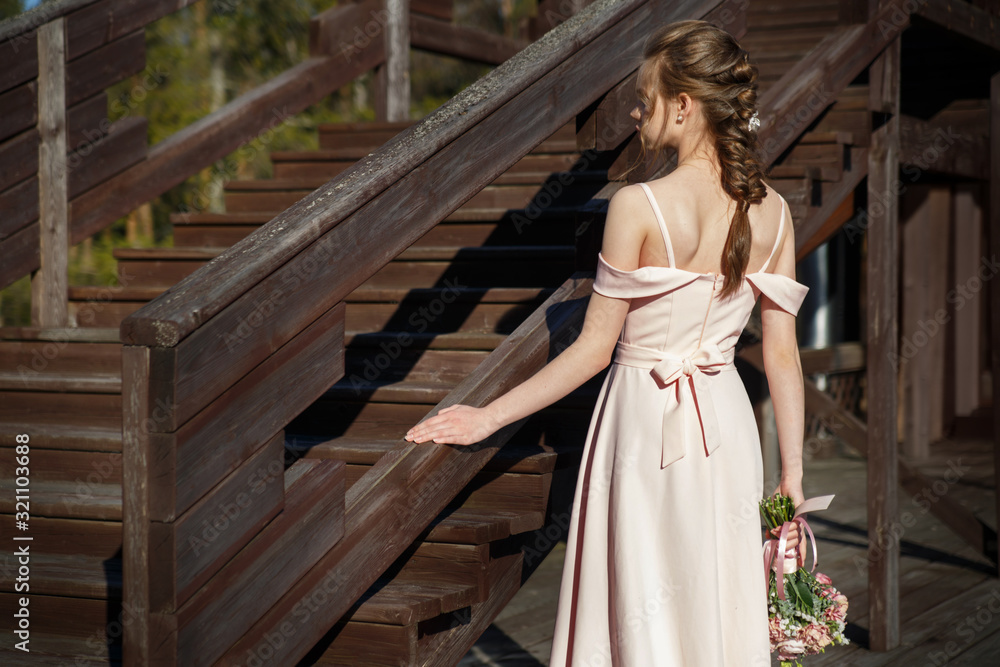 Portrait of a young bride girl in a delicate pink dress with a bouquet in her hands on a rustic terrace in summer day view from the back.