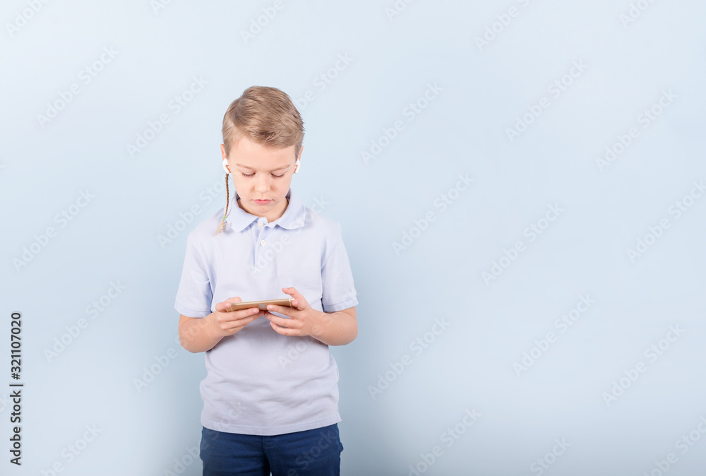 a boy plays a smart phone in wireless headphones on a blue background, concept of children gadgets addiction a boy plays a smart phone in wireless headphones on a blue background, concept of children gadgets addiction