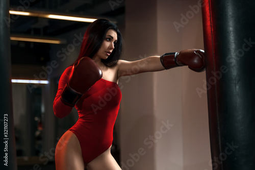 Sexy fitness brunette woman in red body hits sandbag with boxing gloves in gym © Peterfilm