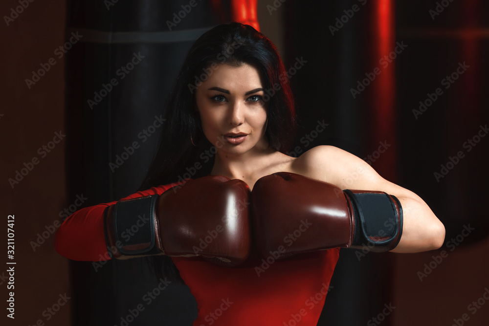 Sexy fitness brunette woman in red body hits sandbag with boxing gloves in gym