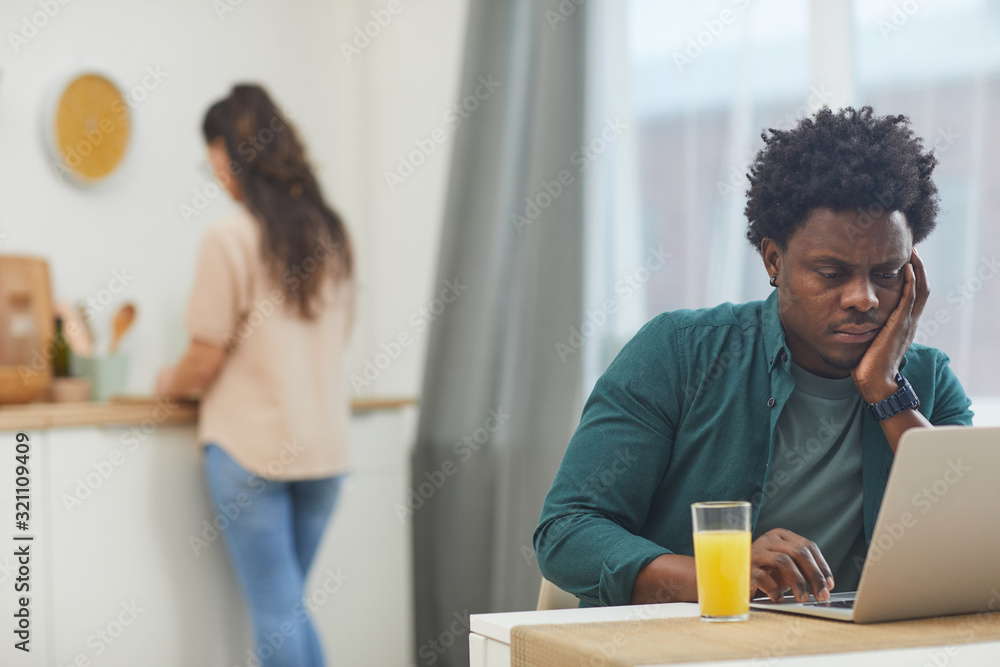 African serious man sitting at the table with glass of juice and working on laptop with his girlfriend cooking in the background in the kitchen African serious man sitting at the table with glass of juice and working on laptop with his girlfriend cooking in the background in the kitchen