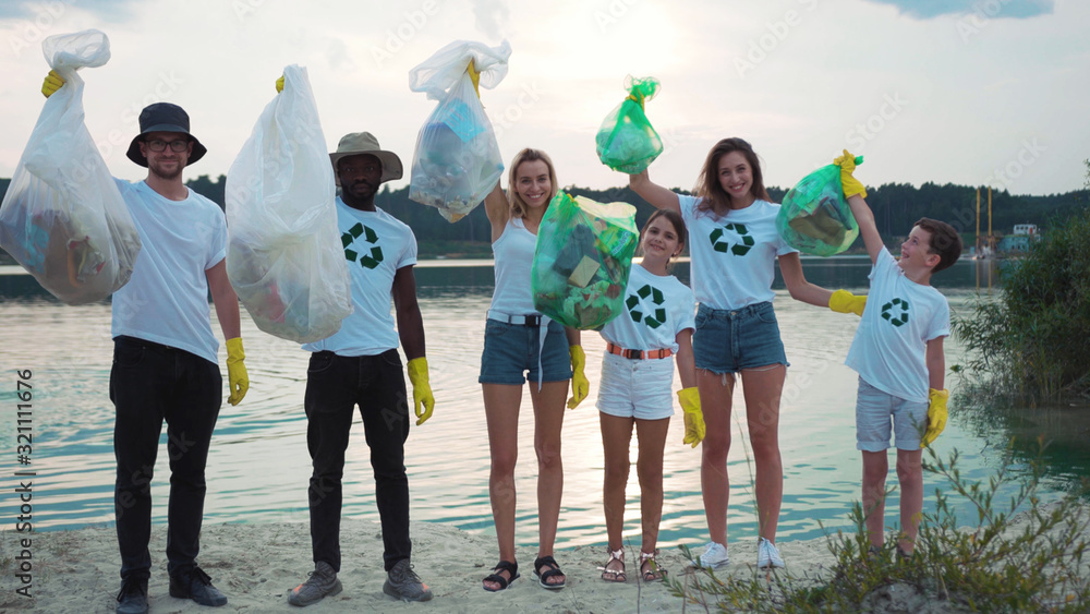 Obraz premium Team of multi-race happy ecology volunteers smiling posing on sandy beach at beautiful lake demonstrating trash bags. Successful nature cleanup. Fight pollution.