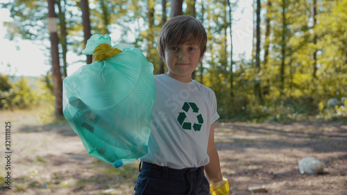 Child boy voluteers activists in gloves tidying up rubbish in park or forest look at camera smile save environmentplastic pollution bag bottle recycle ecology garbage nature care slow motion photo