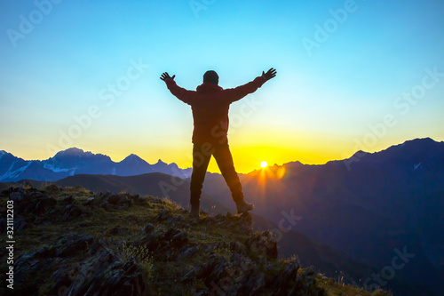 man with raised hands standing on the mountain face to sunrise. Tourist traveler in the mountains meets a sunny dawn.