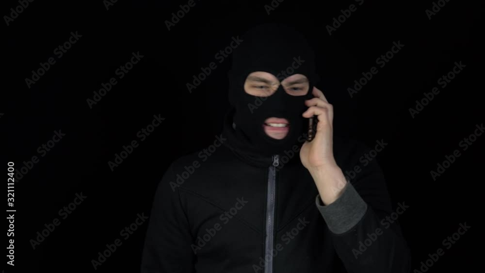 A man in a balaclava mask is talking on a phone. A thug screaming on the phone on a black background. A man in a balaclava mask is talking on a phone. A thug screaming on the phone on a black background.
