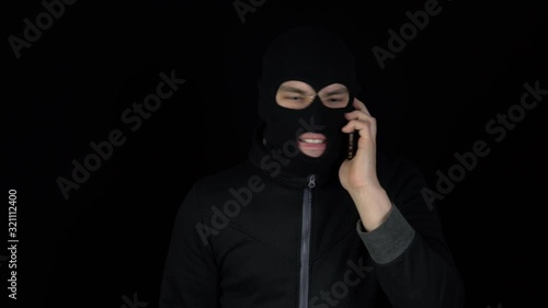 A man in a balaclava mask is talking on a phone. A thug screaming on the phone on a black background.