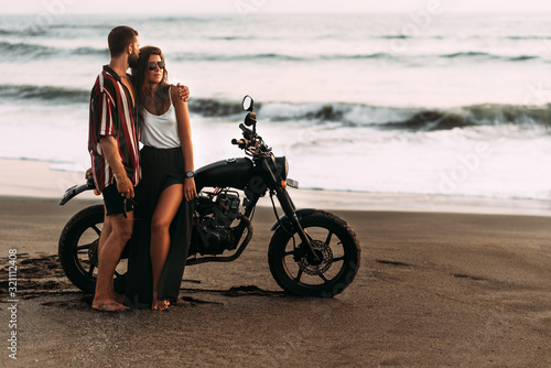 Couple on a motorcycle on the beach. A couple in love on the beach meets the sunset. A man embraces a woman on the beach. Beautiful couple on a motorcycle. Travel on a motorcycle. Lovers on the beach