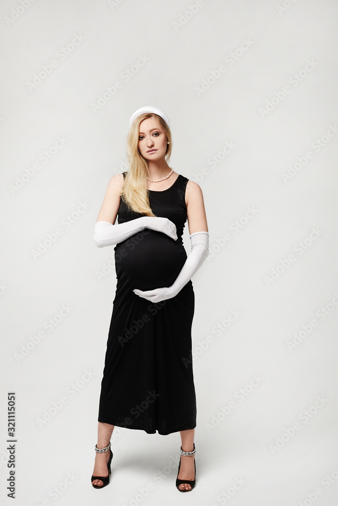 Beautiful pregnant woman in black evening dress and long white gloves touching her belly and posing on a white background, isolated. High fashioned portrait of young mother anticipation of the baby. Beautiful pregnant woman in black evening dress and long white gloves touching her belly and posing on a white background, isolated. High fashioned portrait of young mother anticipation of the baby.