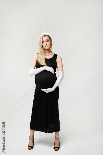 Beautiful pregnant woman in black evening dress and long white gloves touching her belly and posing on a white background, isolated. High fashioned portrait of young mother anticipation of the baby.