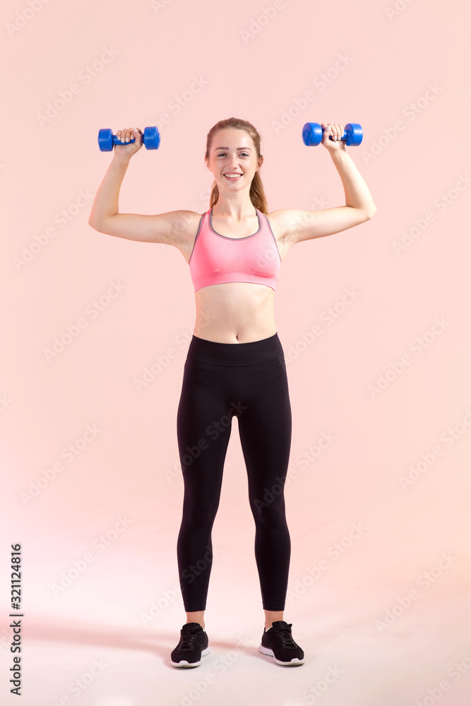 Women's Fitness. Young sports woman with dumbbells in hands standing on a light pink background. Women's Fitness. Young sports woman with dumbbells in hands standing on a light pink background.