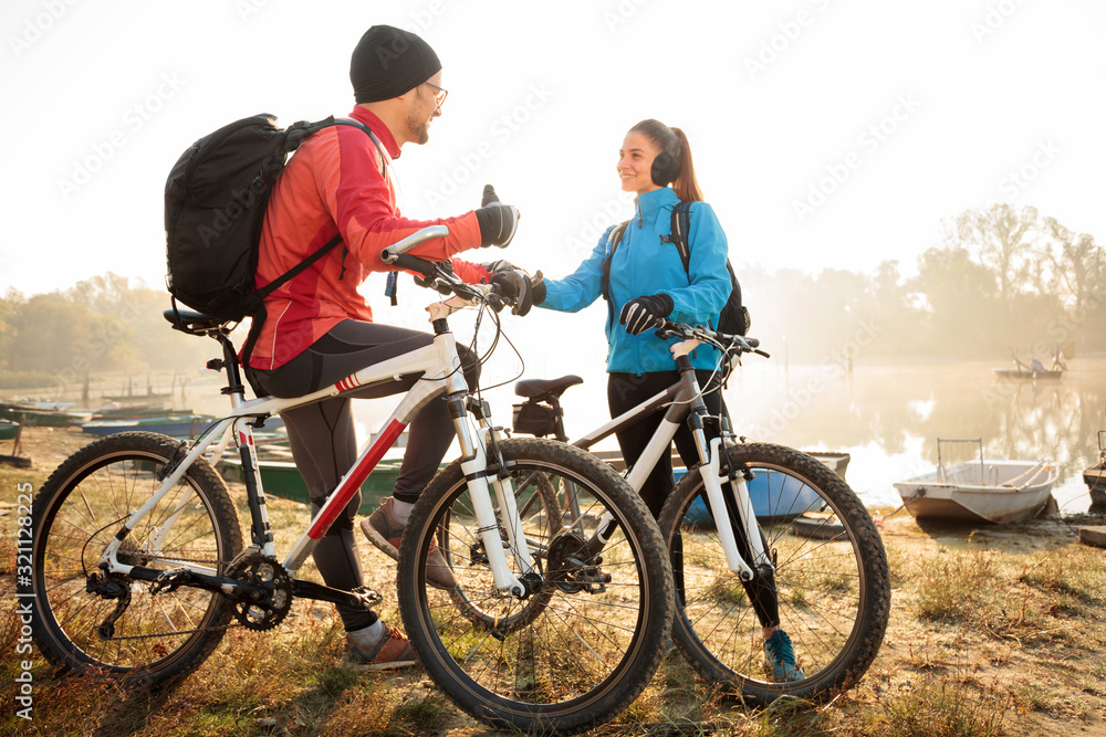 Beautiful happy young active couple enjoying early morning mountain bike ride, resting and talking by the lake. Sun rising through the fog above water in the background.