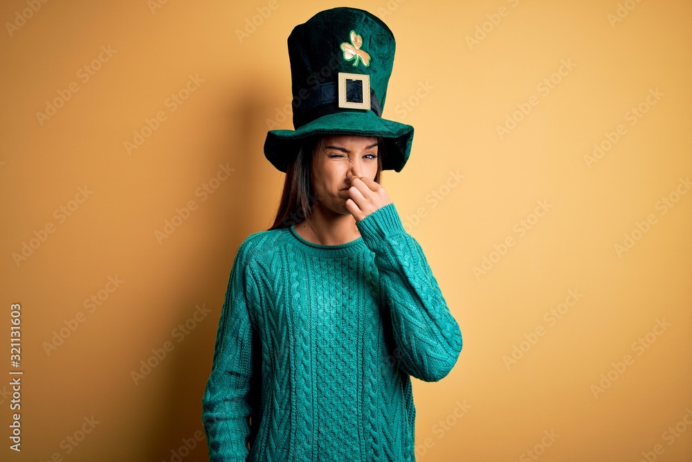 Young beautiful brunette woman wearing green hat with clover celebrating saint patricks day smelling something stinky and disgusting, intolerable smell, holding breath with fingers on nose. Bad smell Young beautiful brunette woman wearing green hat with clover celebrating saint patricks day smelling something stinky and disgusting, intolerable smell, holding breath with fingers on nose. Bad smell