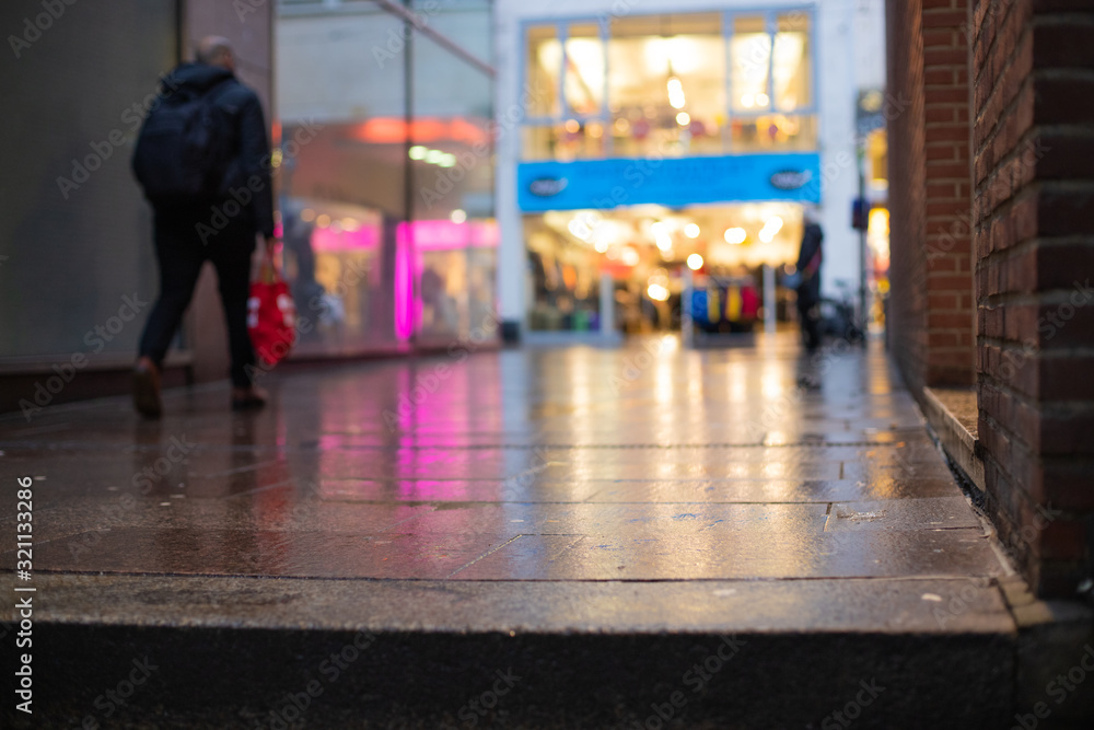 Out of focus picture of a man shopping in the city centre with reflections of lights and store fronts on a blurry wet pavement