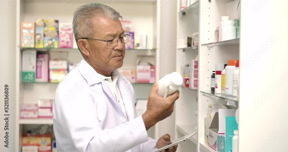 Asian senior pharmacist holding computer tablet using for checking medication details on a box in pharmacy drugstore. Asian senior pharmacist holding computer tablet using for checking medication details on a box in pharmacy drugstore.