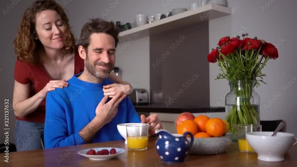 Handheld shot of happy woman embracing and kissing man sitting at table eating breakfast in kitchen Handheld shot of happy woman embracing and kissing man sitting at table eating breakfast in kitchen