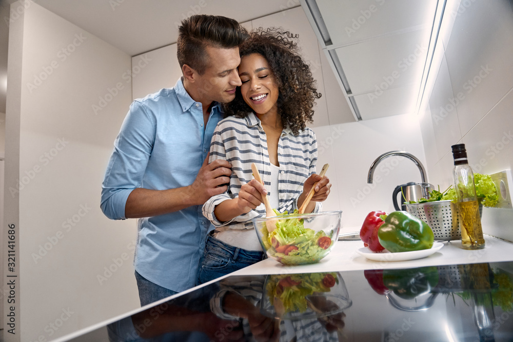 Romantic Date. Young multiethnic couple standing at kitchen cooking dinner husband hugging and kissing wife mixing salad smiling delightful
