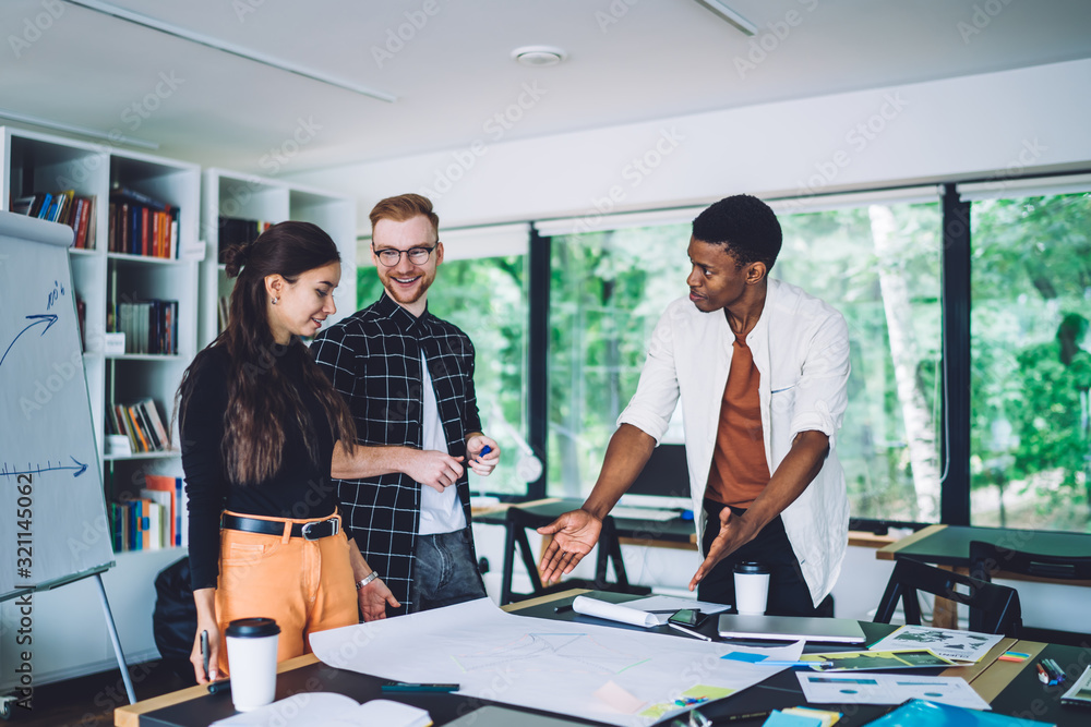 Cheerful adult multiethnic colleagues examining information in blueprint in modern office Cheerful adult multiethnic colleagues examining information in blueprint in modern office