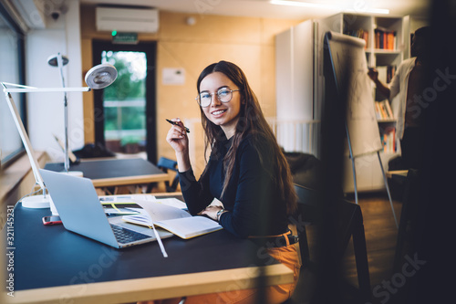 Portrait of successful hipster girl in optical glasses for vision protection smiling at camera during time for e learning on laptop computer, happy female freelancer doing distance job in coworking