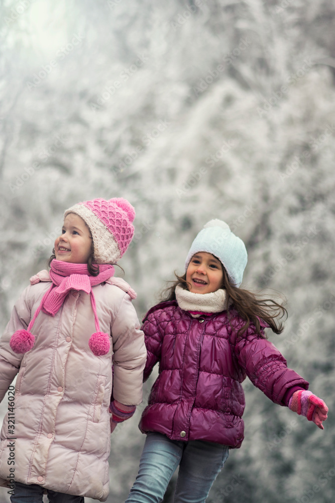 Two sweet little sisters walking between frozen trees during the winter cold day. They enjoying at the wintertime.