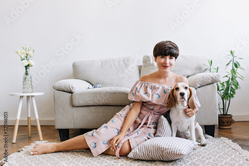 Blissful brunette girl in cute outfit sits on carpet in front of gray sofa with her puppy. Indoor portrait of smiling dark-haired lady and white beagle dog with brown ears posing in room with plant.