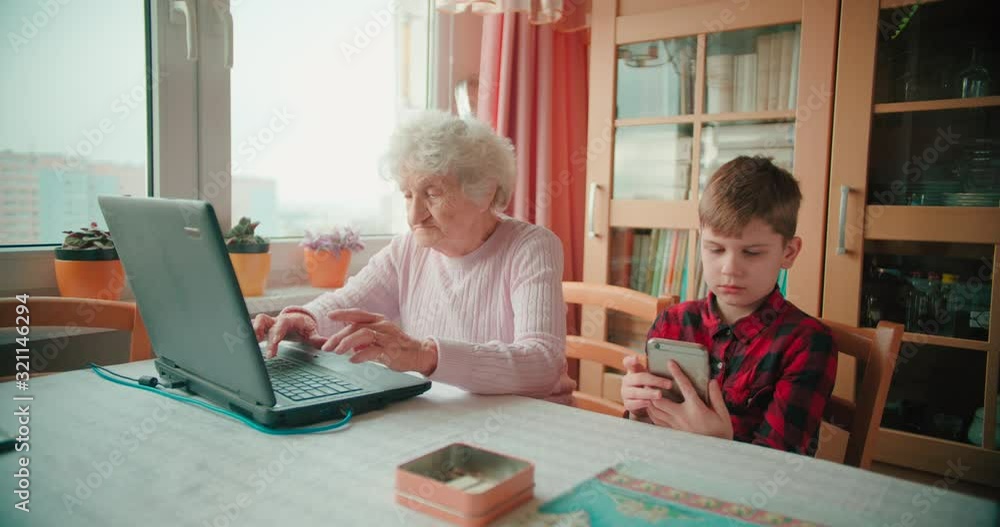 Grandmother typing on laptop keyboard while grandson plays with smartphone Grandmother typing on laptop keyboard while grandson plays with smartphone