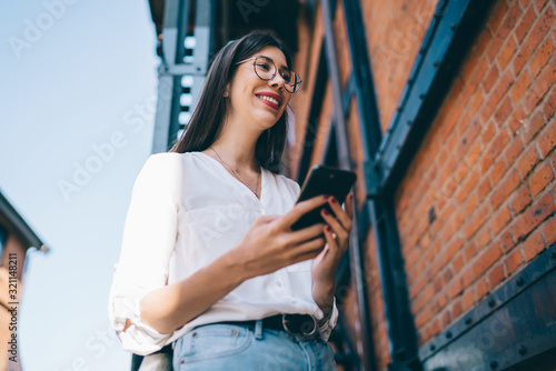 Below view of happy cheerful content blogger holding modern digital gadget for making mobile publication to social networks, smiling Hispanic millennial in optical spectacles using 4g on cellular