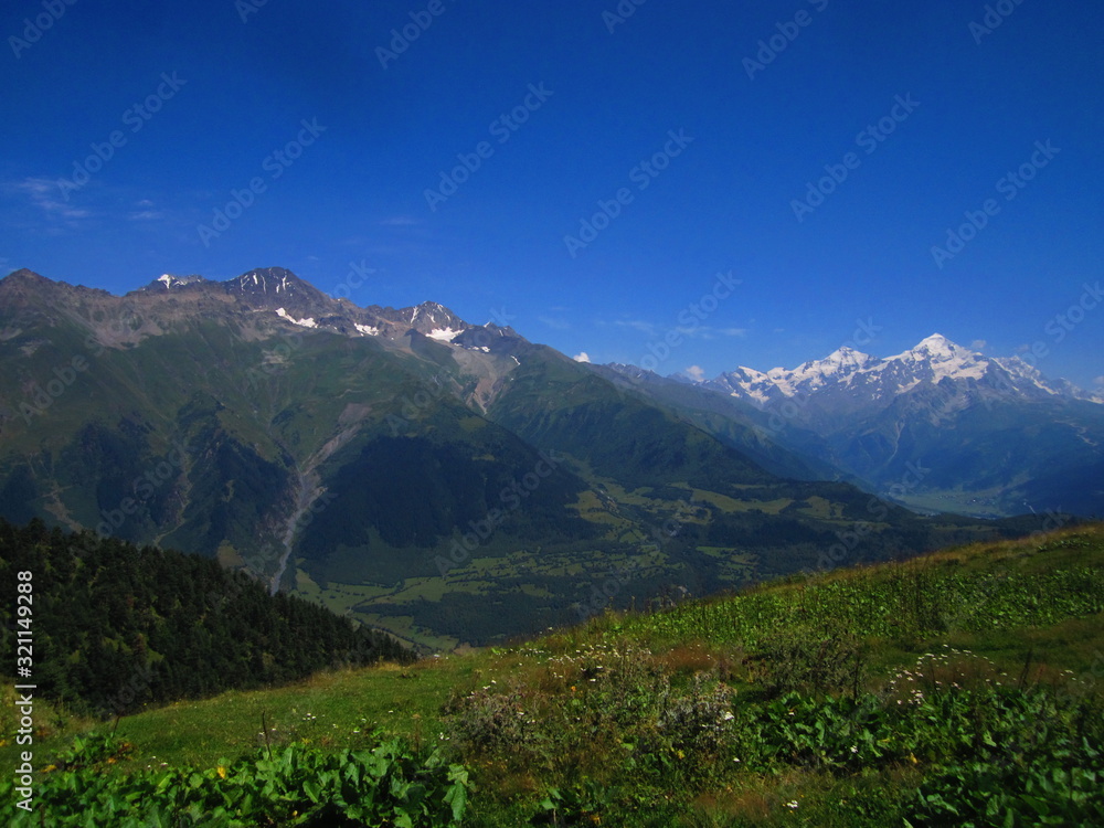 Mountain landscape of Svaneti on bright summer sunny day. Mountain lake, hills covered green grass on snowy rocky mountains background. Caucasus peaks in Georgia. Amazing view on wild georgian nature