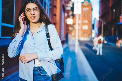 Young woman in spectacles walking on city street talking on mobile phone in roaming, positive casually dressed female satisfied with connection having conversation while strolling outdoors