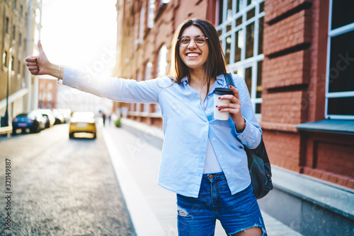 Cheerful caucasian female laughing enjoying travel in city gesture stopping car on street with coffee to go, cheerful woman traveler enjoying visiting town waving and waiting for cab on sunny day