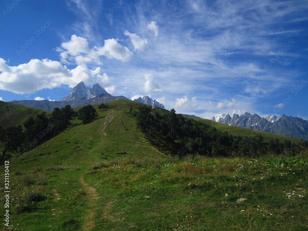 Fototapeta premium Mountain landscape of Svaneti on bright summer sunny day. Mountain lake, hills covered green grass on snowy rocky mountains background. Caucasus peaks in Georgia. Amazing view on wild georgian nature