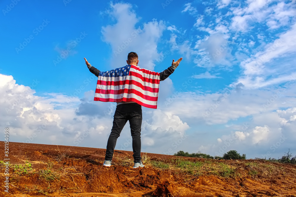 American, a patriot of the country, with the USA flag on his shoulders raised his hands to the blue sky with clouds on top of the mountain. Photos on the theme: love of America, Independence Day