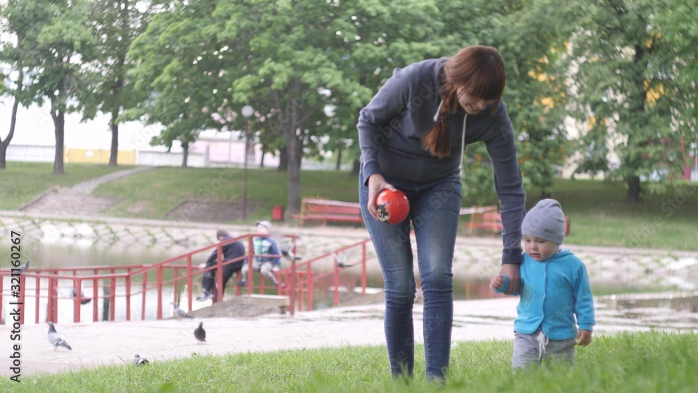 Young woman with her little baby son are walking in the city park in a summer cloudy day with pond and trees on the background in 4K slow motion video. Young woman with her little baby son are walking in the city park in a summer cloudy day with pond and trees on the background in 4K slow motion video.