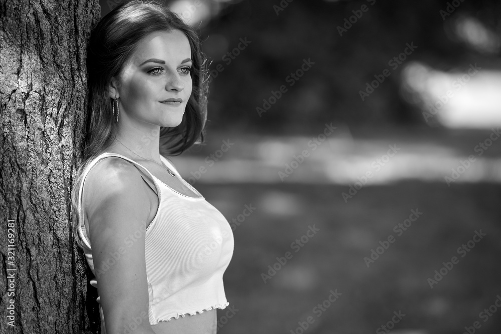 Black and white portrait of beautiful young woman in white tank top leaning against tree - subtle highlights and shadows Black and white portrait of beautiful young woman in white tank top leaning against tree - subtle highlights and shadows