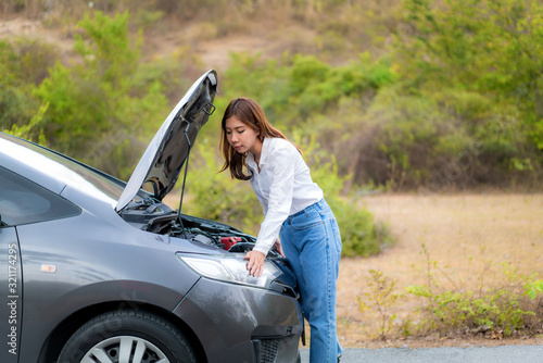 Asian young unhappy woman inspecting broken car engine in front of the open hood broken down car On Country Road Waiting for road assistance service. Broken Car On The Road in During journey..