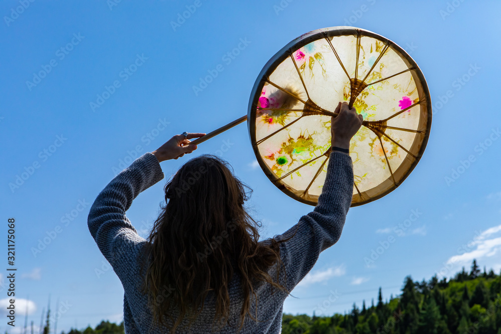 Fototapeta premium Rear closeup view of young shamanic woman holding sacred native frame drum with raised hands worshiping in Northern Quebec in Canada
