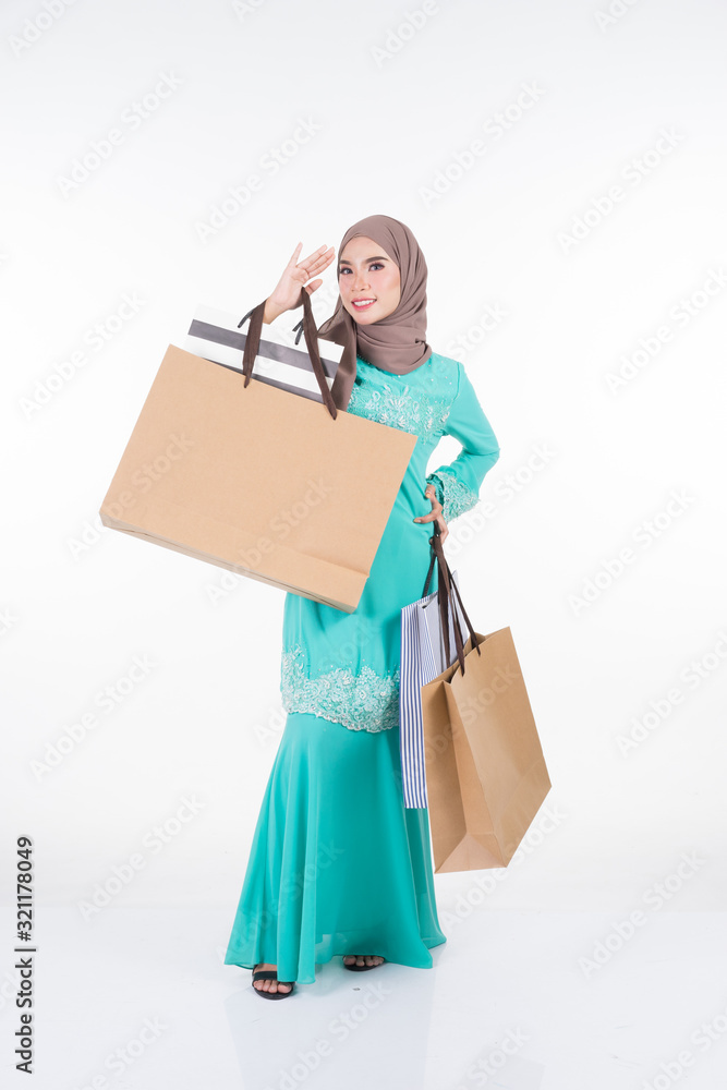 A beautiful and excited Muslim female model in a Asian traditional dress modern kurung carrying shopping bags isolated on white background. Eidul fitri festive preparation shopping concept. A beautiful and excited Muslim female model in a Asian traditional dress modern kurung carrying shopping bags isolated on white background. Eidul fitri festive preparation shopping concept.