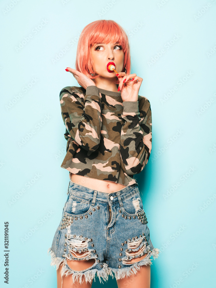 Portrait of young beautiful hipster bad girl in trendy jeans shorts and earring in her nose.Sexy carefree smiling woman in pink wig posing in studio.Positive model licking round sugar candy Portrait of young beautiful hipster bad girl in trendy jeans shorts and earring in her nose.Sexy carefree smiling woman in pink wig posing in studio.Positive model licking round sugar candy