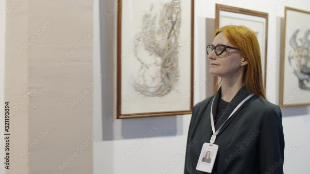 Beautiful redhead woman with visitor badge on lanyard around neck walking in art gallery, looking at artworks on the wall and then posing for camera and smiling