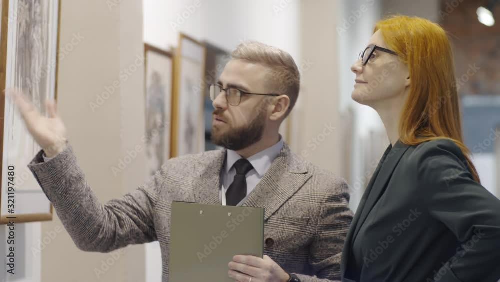 Experienced male gallery docent with clipboard telling about artwork to redhead woman while presenting modern art collection to her Experienced male gallery docent with clipboard telling about artwork to redhead woman while presenting modern art collection to her