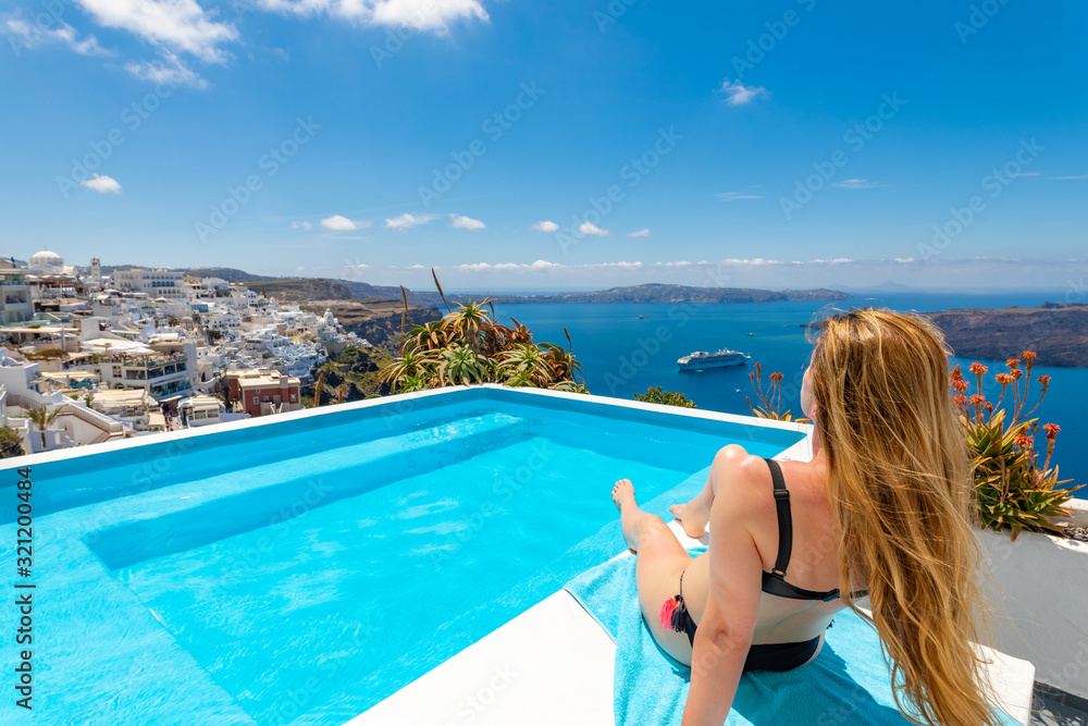 Beautiful young blonde woman in a tan color swimsuit lying by the pool on the island of Santorini in Greece, in the background is a beautiful amazing landscape on the mountains and the sea