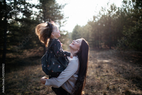 Young beautiful mom turns over daughter in her arms on the forest background on sunset. Laughing mom and daughter. Parenthood. Mom hugs daughter