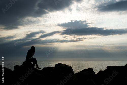 silhouette of a girl looking towards the horizon, sitting on the seashore stones