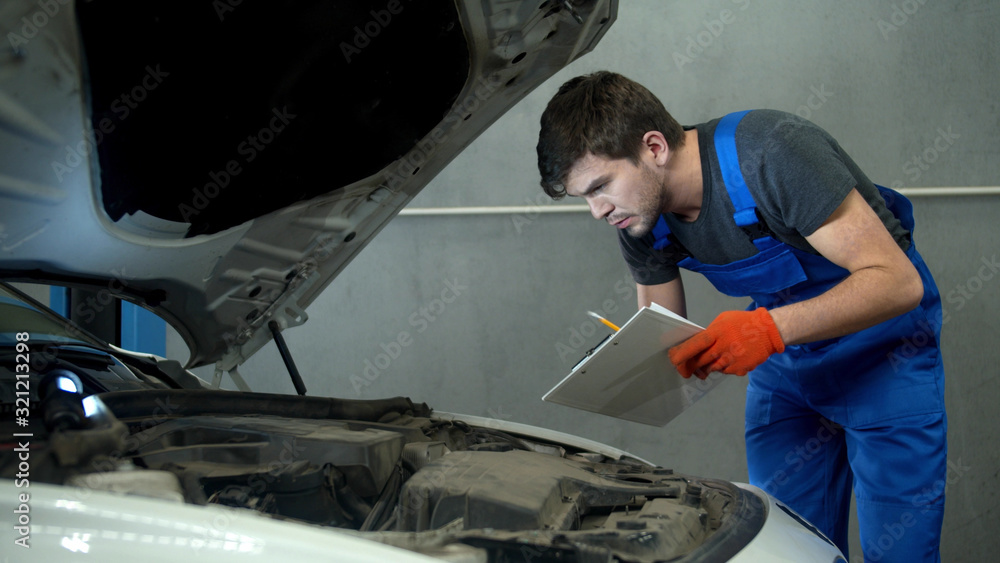 Mechanic examines the motor of car and takes notes Mechanic examines the motor of car and takes notes