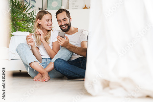 Loving couple sit on floor indoors using mobile phone.